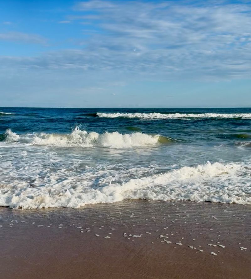 A close view of ocean waves rolling toward the shore, white foam meeting wet sand under a soft blue sky. The light catches the water’s surface, capturing both motion and stillness — like sound before it becomes language.