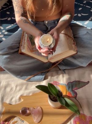Hands holding a candle over an open Bible with journal pages, surrounded by warm light and a table with a plant and sacred objects — a quiet moment of resurrection embodiment.